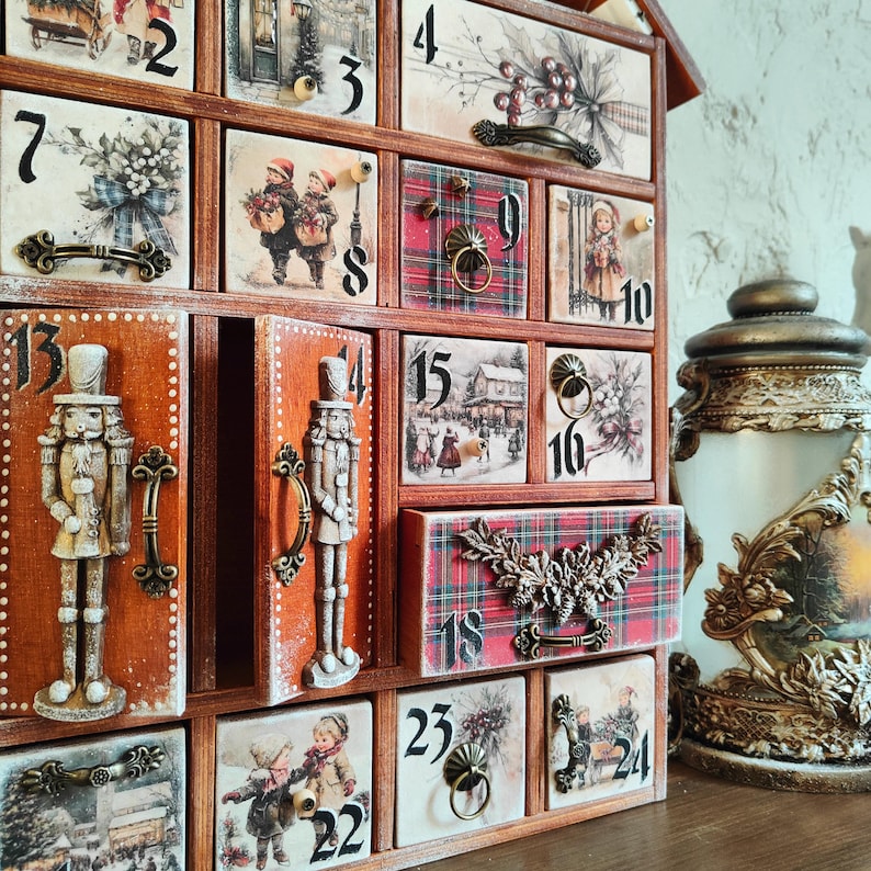 May include: A wooden advent calendar with twenty-four numbered drawers featuring various holiday illustrations, including nutcrackers, winter scenes, and plaid patterns. Each drawer has a metal handle or pull, and the unit is finished in a warm brown tone.