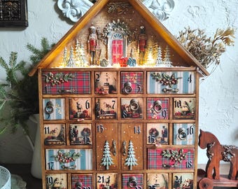 May include: A wooden advent calendar shaped like a house with twenty-four numbered drawers. The top features a miniature scene with two nutcrackers, a red door, and illuminated trees. Each drawer displays festive patterns, holly, and vintage imagery.