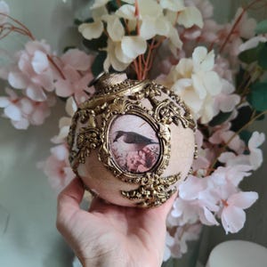 Pode incluir: Enfeite ornamentado creme com uma ilustração de pássaro em uma moldura dourada. O enfeite apresenta filigranas douradas e um acabamento craquelado. Uma peça decorativa para as festas ou decoração de casa.