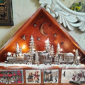 May include: A wooden advent calendar with a triangular top featuring a train scene with snow-dusted trees, a crescent moon, and stars illuminated by string lights. The base has four drawers decorated with winter illustrations and the number 4.