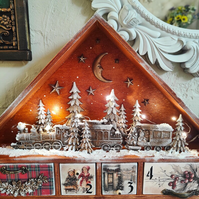 May include: A wooden advent calendar with a triangular top featuring a train scene with snow-dusted trees, a crescent moon, and stars illuminated by string lights. The base has four drawers decorated with winter illustrations and the number 4.