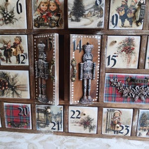 May include: A wooden advent calendar with twenty-four numbered drawers featuring vintage-style Christmas illustrations, including children, winter landscapes, and holly. Two central drawers are decorated with detailed nutcracker figures.