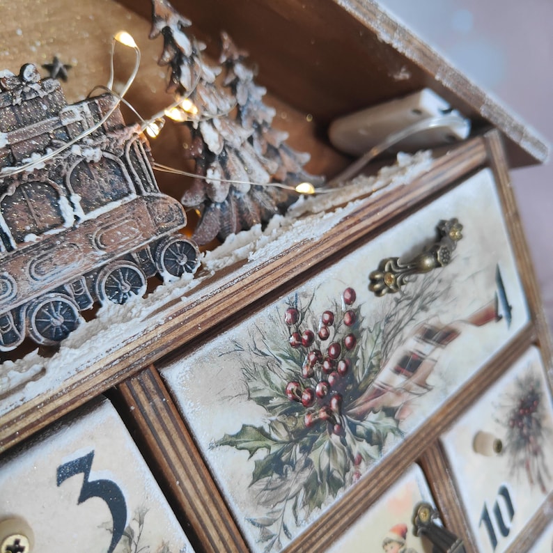 May include: A wooden advent calendar with numbered drawers featuring holly and berry illustrations. The top section displays a decorative train and pine tree cutout with integrated warm white fairy lights and faux snow accents.