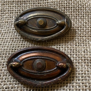 May include: Two antique oval cabinet door pulls with a decorative design. The top pull is brass and the bottom pull is copper.