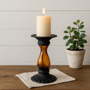 May include: A lit ivory candle sits atop a black metal and amber glass candlestick holder. The candlestick has a decorative base and a flared top. A small potted plant is in the background.