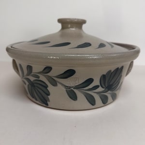 May include: A beige stoneware casserole dish with a lid. The dish features a floral design in dark blue. The lid has a small handle and a similar dark blue leaf pattern. The dish has two small handles on the sides.