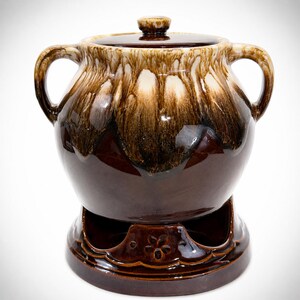 May include: A brown and cream glazed ceramic pot with two side handles and a matching lid. The pot sits on a circular base featuring a cutout design with a small embossed flower detail. The glaze features a drip pattern from the rim.