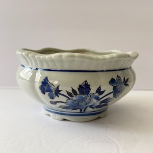May include: A small, off-white ceramic planter with a scalloped rim and a blue floral design. The planter has a rounded shape and a footed base. The blue floral pattern includes flowers and leaves. The planter is suitable for small plants.