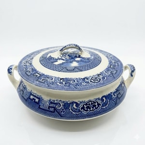 May include: A blue and white ceramic serving bowl with a lid. The bowl features a detailed pattern of landscapes and geometric designs. It has two handles and a knob on the lid. The color scheme is primarily blue on a white background.