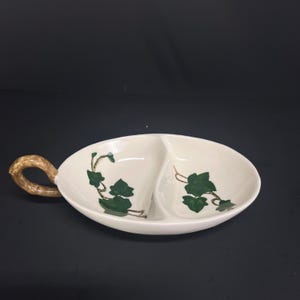 May include: Cream-colored, divided serving dish with a handle. The dish features green ivy leaf designs and a textured, brown handle. The oval dish is divided into two sections, perfect for serving snacks or appetizers.
