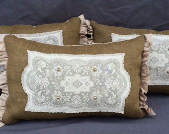 May include: Four decorative throw pillows. Each pillow has a brown burlap base with a white lace appliqué and gold-colored button accents. The pillows are rectangular with ruffled edges in a light tan color.
