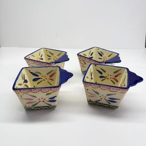 May include: Four square ceramic bowls with a cream-colored base, adorned with colorful floral patterns and a blue rim and handle. The bowls have a decorative pink and blue border.