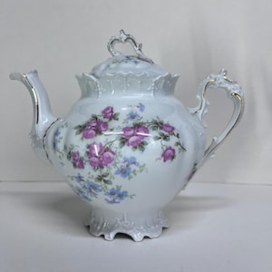 May include: White porcelain teapot with a floral design featuring pink roses and blue flowers. The teapot has a decorative handle, spout, and lid with gold trim. A classic piece for tea parties or display.