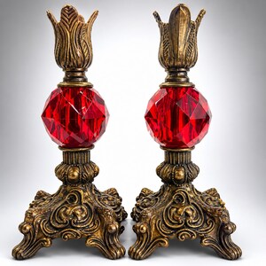 May include: A pair of candle holders featuring ornate, antiqued gold-tone metal bases with intricate scrollwork and leaf-like tops. Each holder is centered with a large, faceted, vibrant red glass sphere that catches the light, creating a striking contrast between the metallic finish and the deep red glass elements.