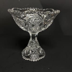May include: A clear, ornate glass pedestal bowl with a scalloped rim. The bowl features intricate patterns and designs throughout, including starburst and floral motifs. The bowl is elevated on a detailed pedestal base.