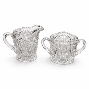 May include: A clear glass creamer and sugar bowl set. The creamer has a spout and handle, while the sugar bowl has two handles. Both pieces feature a detailed, textured pattern.