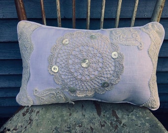 May include: A lavender pillow with a crocheted doily and pearl buttons. The pillow is sitting on a weathered wooden chair.