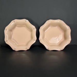 May include: Two light peach-colored ceramic plates with an octagonal shape and scalloped edges. Each plate has small, raised decorative elements around the rim. The plates are set against a dark background.