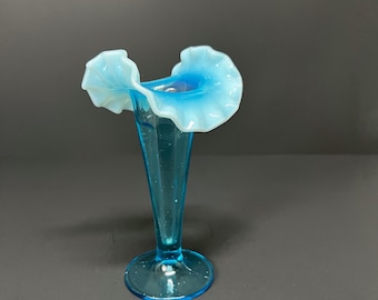 May include: A light blue glass vase with a ruffled rim. The vase has a slender, elongated body and a round base. The ruffled rim is a lighter shade of blue, creating a delicate contrast. The vase is likely vintage.