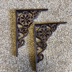 May include: Two ornate cast iron shelf brackets with a brown finish. The brackets have a decorative floral design.