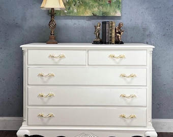 May include: White painted chest of drawers with five drawers and ornate gold handles. The chest has a scalloped base and sits on four cabriole legs.