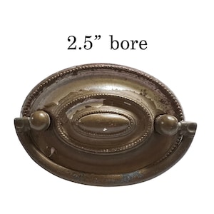 May include: An antique brass drawer pull with an oval center and beaded trim. The pull has a 2.5" bore and two rounded end pieces. The metal shows signs of wear and age.