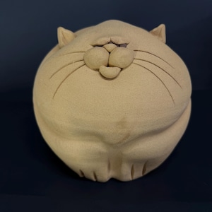 May include: A beige ceramic cat figurine with a round body and a smiling face. The cat has small, triangular ears and long whiskers. The figurine is a decorative item.