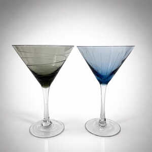 May include: Two martini glasses with clear stems and bases. The left glass is smoke-gray with thin, etched horizontal spiral lines. The right glass is blue with vertical etched lines radiating from the base of the bowl toward the rim.