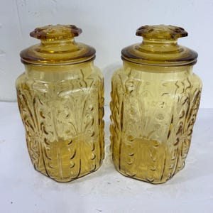 May include: Two amber-colored glass canisters with matching lids. Each jar features a textured, raised botanical pattern on the exterior surface. The glass is translucent, allowing light to pass through the warm, golden-toned material.