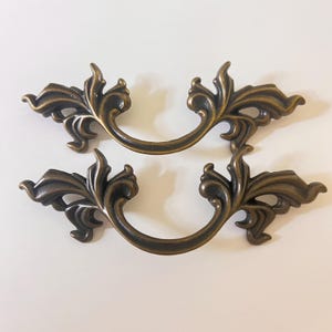 May include: Two antique bronze-colored drawer pulls with a decorative, ornate leaf design. Each pull features a curved handle and detailed floral accents, perfect for adding a vintage touch to furniture. The pulls are approximately 6 inches wide.