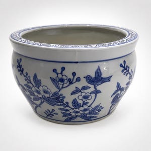 May include: A round ceramic bowl with a white base and blue painted details. The exterior features a pattern of birds perched on flowering branches. The rim is decorated with a continuous blue scroll design. The interior is plain white.