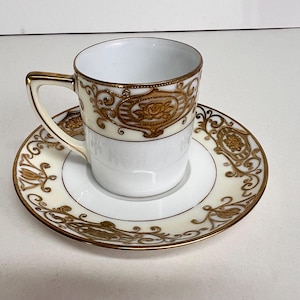 May include: A white teacup and saucer set with gold filigree and floral designs. The cup has a gold handle and a band of gold detailing around the rim. The saucer mirrors the cup's design, with gold accents and a delicate pattern.