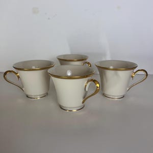 May include: Four off-white teacups with gold trim and handles. The cups have a classic design, with a slightly flared shape and a rounded handle. The gold trim accents the rim and handle, adding a touch of elegance to the set.