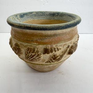May include: A weathered, beige ceramic planter with a dark blue rim. The planter features a band of reddish-brown and is adorned with embossed seashell designs around the middle. The pot is suitable for indoor or outdoor use.