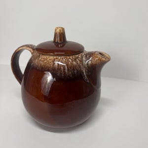 May include: A brown ceramic teapot with a lid and handle. The teapot has a glossy finish with a two-tone design, featuring a darker brown base and a lighter, speckled brown top. The spout is on the right side.