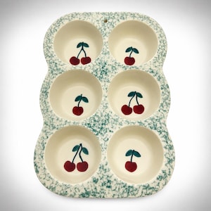 May include: A ceramic muffin pan with six circular cups, featuring a cream-colored interior and a speckled teal and white exterior. Each cup is decorated with a hand-painted illustration of two red cherries with green stems at the bottom.