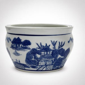 May include: A white ceramic bowl featuring a blue transferware pattern depicting a landscape with pagodas, trees, and a small boat on water. The bowl has a wide, rounded body with a smooth rim and a solid blue line encircling the top edge.