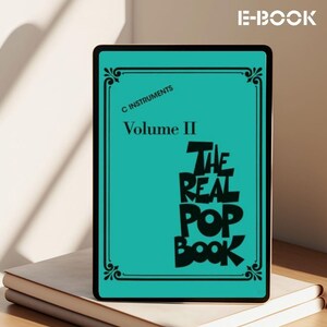 May include: An e-book titled "The Real Pop Book, Volume II" displayed on a tablet. The tablet has a teal background with black text and decorative borders. The tablet rests on top of two stacked, off-white books.