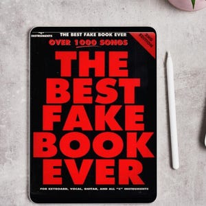 May include: A black tablet displaying "THE BEST FAKE BOOK EVER" in large red letters. The text also includes "OVER 1000 SONGS" and "FOR KEYBOARD, VOCAL, GUITAR, AND ALL 'C' INSTRUMENTS". A white stylus and a small potted plant are also visible.