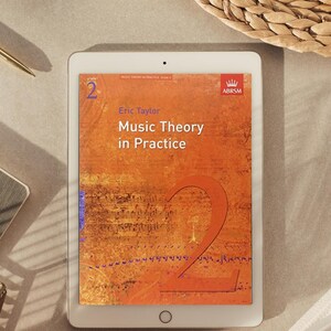 Puede incluir: Una tableta blanca que muestra la portada del libro Music Theory in Practice Grade 2 de Eric Taylor. La portada es naranja con un número 2 grande, notación musical y el logotipo de ABRSM en la esquina superior derecha. La tableta tiene una cámara.