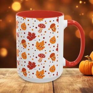 Könnte beinhalten: Weiße Keramiktasse mit rotem Innenraum und Griff. Die Tasse hat ein sich wiederholendes Muster aus Cartoon-Blättern, Eicheln und Pilzen in Orange-, Rot- und Brauntönen. Die Tasse steht auf einer Holzoberfläche.