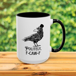 May include: White ceramic mug with a black interior and handle. The mug features a black and white illustration of a crow wearing a top hat and glasses. The text "Politely, F-CAW-F" is below the crow.