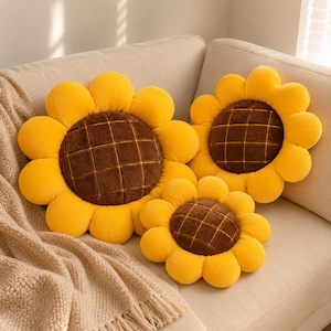 May include: Three round throw pillows shaped like sunflowers. Each pillow features bright yellow corduroy petals surrounding a dark brown, plush, grid-patterned center. The pillows vary in size and are arranged together on a neutral surface.