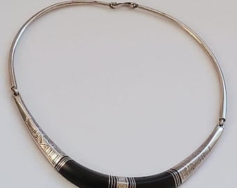 May include: A silver and black choker necklace with an engraved design. The necklace is made of a thin silver band with a black resin insert. The insert has a geometric pattern and is accented with silver trim.