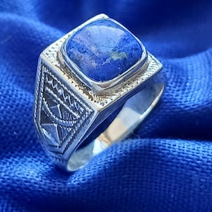 Op de afbeelding: Een zilveren ring met een vierkante, blauwe edelsteen. De ring heeft gedetailleerde geometrische patronen aan de zijkanten en een getextureerde zetting rond de steen. De ring is geplaatst tegen een blauwe achtergrond.