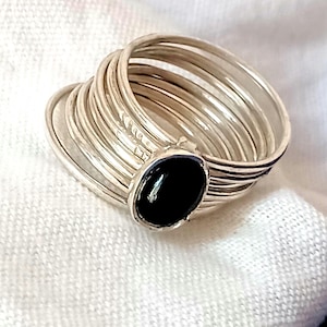 Peut inclure: Une bague multi-anneaux avec une finition argentée polie et une pierre noire de forme ovale sertie dans une monture en argent. Le design se compose de plusieurs anneaux fins et empilés, réunis à la base de la pierre noire centrale.