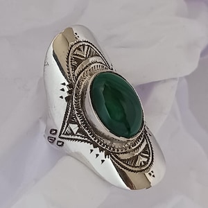 Peut inclure: Une bague en argent ornée d'une pierre ovale vert foncé. La bague présente des motifs gravés complexes autour de la pierre et sur le corps. L'ensemble est mis en valeur sur un fond blanc, soulignant ses détails.