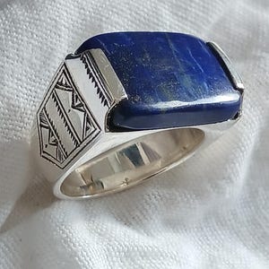 Könnte beinhalten: Ein silberner Ring mit einem großen, rechteckigen, tiefblauen Lapislazuli-Stein. Der Ring hat dekorative geometrische Muster an den Seiten und eine polierte Oberfläche, vor weißem Hintergrund.