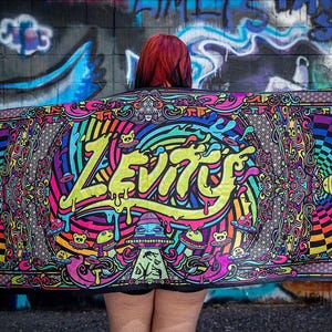 May include: A vibrant tapestry with the word "LEVITY" in dripping yellow letters. The design features swirls, geometric patterns, and cartoonish elements like UFOs and bears. The background is a graffiti-covered wall with colourful artwork.