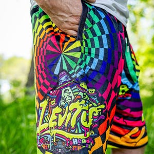 May include: Multi-colored shorts featuring a vibrant checkerboard pattern in shades of neon green, blue, orange, and purple. The design includes a graphic print with the text "LEVITY" in yellow, surrounded by small alien and UFO illustrations.